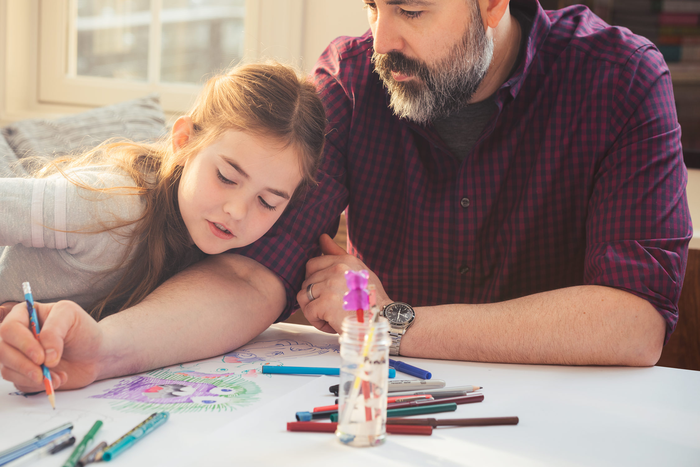 A father teaches his daughter to draw with markers on paper.